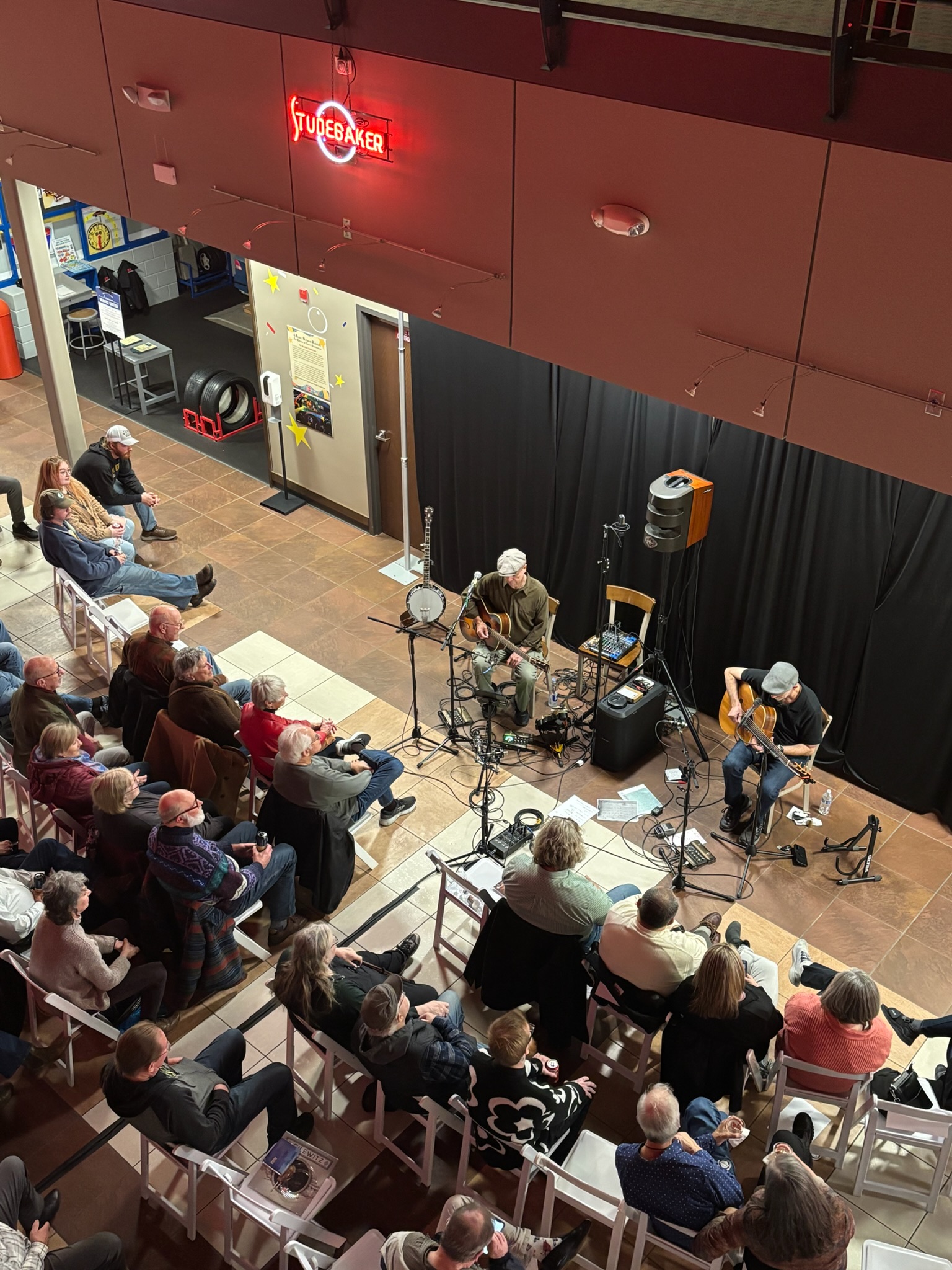 A crowd of people sit on white chairs listening to two men at the front of the room playing guitars.
