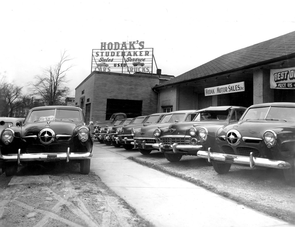 A car dealership with rows of Studebakers from the 1950s. A sign on top of the build says Hodak's Studebaker