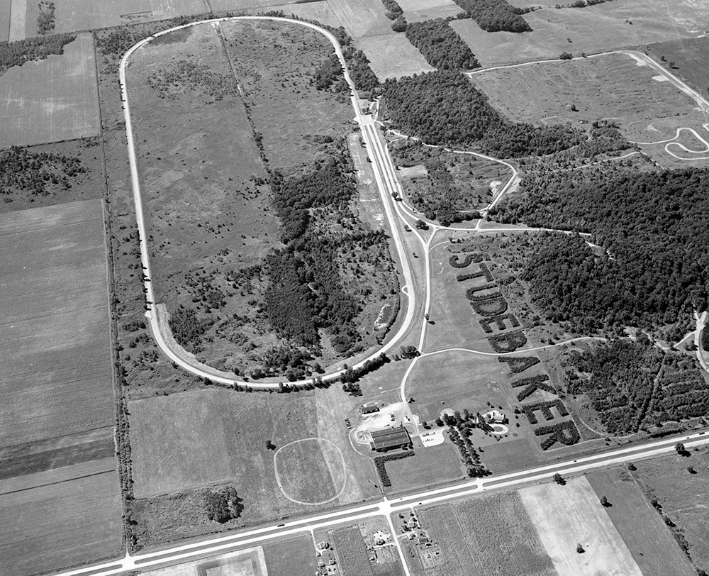 A testing racetrack is seen from an arial view surrounded by fields. On the right side of the image, trees that spell out STUDEBAKER can be seen.