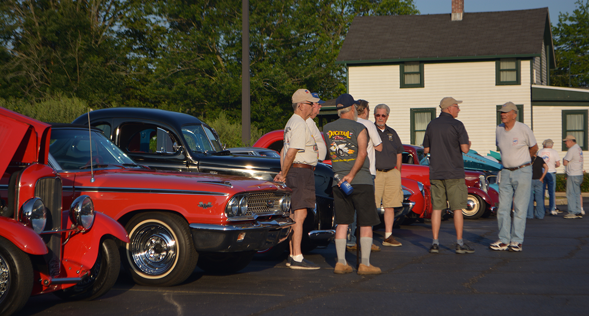 Men stand around hot rod vehicles in a parking lot.